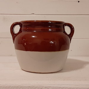 May include: A brown and white ceramic crock with two handles. The crock is glazed and has a rustic look.