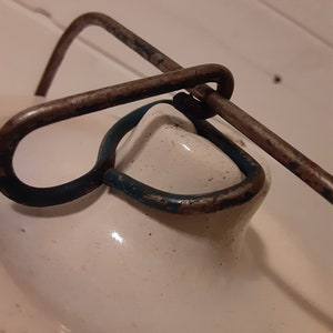 Puede incluir: Primer plano de una tapa de cer&aacute;mica blanca con un asa de metal oxidado. El asa tiene un anillo pintado de azul.