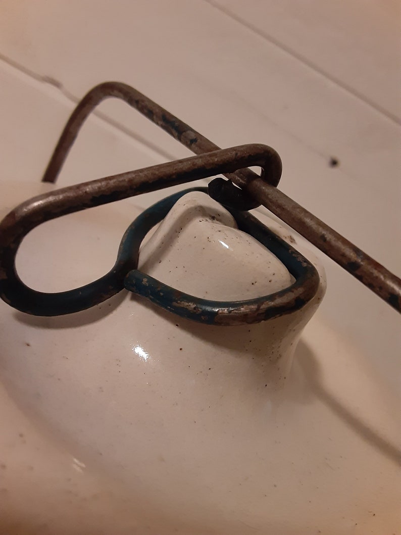 Puede incluir: Primer plano de una tapa de cer&aacute;mica blanca con un asa de metal oxidado. El asa tiene un anillo pintado de azul.