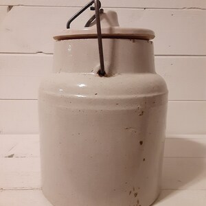 Puede incluir: Un tarro de cer&aacute;mica blanco con un asa de alambre met&aacute;lico y una tapa. El tarro tiene una superficie ligeramente texturizada y parece ser vintage.