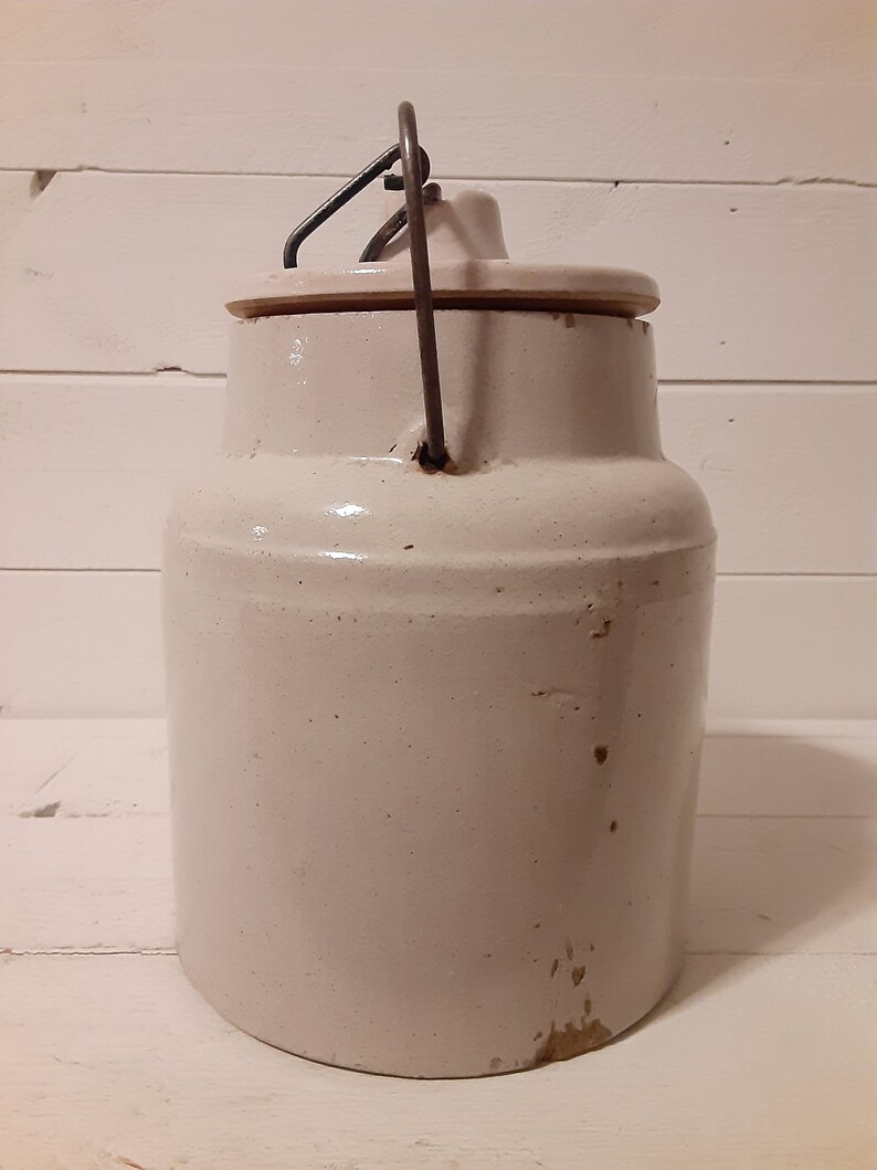 Puede incluir: Un tarro de cer&aacute;mica blanco con un asa de alambre met&aacute;lico y una tapa. El tarro tiene una superficie ligeramente texturizada y parece ser vintage.