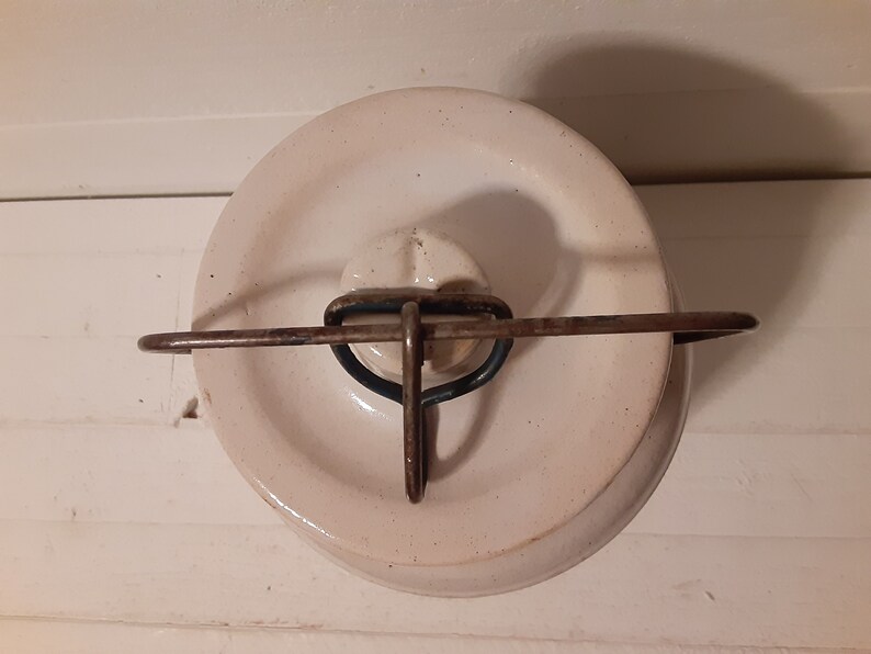 Puede incluir: Tarro de cer&aacute;mica blanco con tapa de metal y asa de alambre. La tapa tiene un pomo en relieve en el centro.