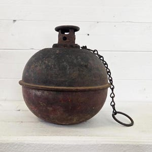 May include: A vintage, spherical metal object with a dark, weathered finish. It features a small, cylindrical top and a chain with a ring. The object has a rusty, aged appearance, suggesting it is an antique.