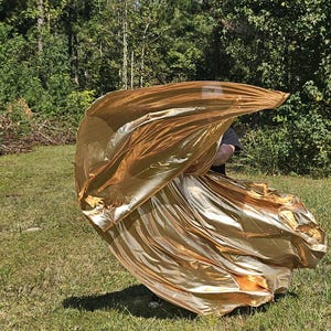 May include: A flowing, golden fabric is held outdoors, creating a dynamic, rippling effect. The material catches the sunlight, displaying a metallic sheen. The background features green grass and trees, suggesting an outdoor setting.