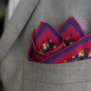 May include: A folded pocket square in a gray suit pocket. The pocket square is primarily fuchsia with a blue border and a geometric pattern of yellow, green, and blue shapes. The suit is a light gray color.