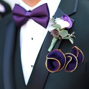 Puede incluir: Una chaqueta de traje azul oscuro con una pajarita morada y un boutonniere de flor blanca. La chaqueta tiene un pa&ntilde;uelo de bolsillo con un dise&ntilde;o de terciopelo morado.