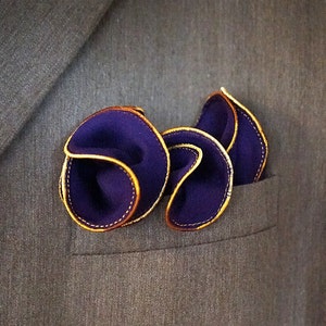 Puede incluir: Un pa&ntilde;uelo de bolsillo de color morado oscuro con ribete dorado, doblado y colocado en el bolsillo de una chaqueta de traje.