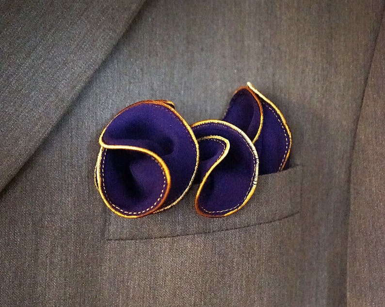 Puede incluir: Un pa&ntilde;uelo de bolsillo de color morado oscuro con ribete dorado, doblado y colocado en el bolsillo de una chaqueta de traje.