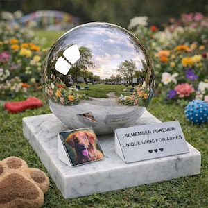 Könnte beinhalten: Eine reflektierende Kugelurne steht auf einem Marmorsockel und spiegelt eine Parkszene wider. Ein kleines Foto eines Hundes und ein Schild mit der Aufschrift "REMEMBER FOREVER UNIQUE URNS FOR ASHES" befinden sich davor. Eine Plüschpfote und ein Hundespielzeug sind im Vordergrund.