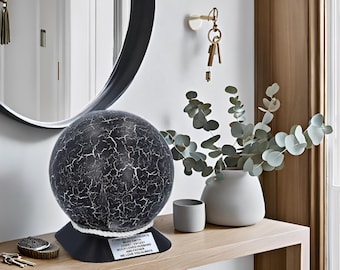 May include: A black and white globe-shaped urn with a rope trim and a plaque that reads "FOR OUR DEAR MARK SMITH JUNE 17, 1967 - JULY 1, 2021 MUCH LOVED HUSBAND AND FATHER WE LOVE YOU ALWAYS".