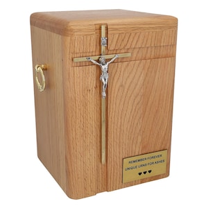 May include: A wooden cremation urn with a gold cross and a silver crucifix. The urn has a brass handle and a plaque that reads "Remember Forever Unique Urns for Ashes".