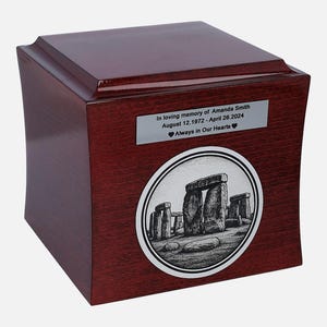Op de afbeelding: Een houten urn met een glanzende, roodbruine afwerking. Een zilveren plaatje toont de inscriptie "In loving memory of Amanda Smith August 12.1972 - April 26.2024 Always in Our Hearts." Een cirkelvormig embleem toont Stonehenge in zwart-wit.