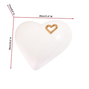 May include: A white heart-shaped decorative object, 11.4 inches wide, 10.2 inches high, and 6.6 inches deep, with a gold heart-shaped accent. The glossy surface reflects light, highlighting the smooth curves of the heart.
