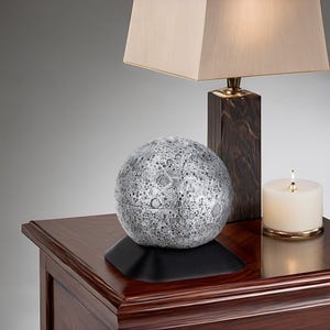 Puede incluir: Una lámpara de plata con forma de luna con una textura de superficie detallada se encuentra sobre una mesa de madera oscura junto a una lámpara con una pantalla beige y una vela.