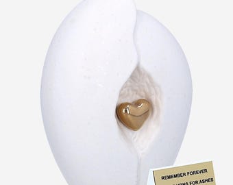 May include: White ceramic urn shaped like an egg with a gold heart inside. The urn has a small opening on the side. The urn is sitting on a gold card that says "Remember Forever Unique Urns for Ashes".