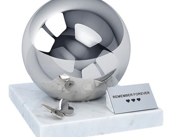 May include: A silver sphere urn with a marble base and two silver butterfly accents. The urn has a small plaque with the text "REMEMBER FOREVER" and three heart symbols.