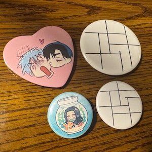 May include: Four colorful pinback buttons on a wooden surface. One pink heart-shaped button features anime characters. Two white oval buttons have a black geometric design. A blue button shows an anime character in a jar with a goldfish.