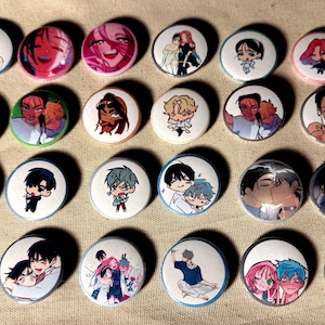 May include: A collection of round, white anime-style button pins. Each pin features a different colorful illustration of anime characters. The pins are arranged on a beige fabric surface.
