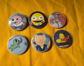 May include: Six round pinback buttons with cartoon character designs. The buttons feature various characters in bright colors, including a pink creature, a yellow face, and a red figure. The buttons are on a yellow background.