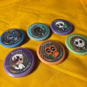 May include: A collection of seven colorful round buttons with cartoon character designs. Each button features a different character name and the text "Dandy's World". The buttons are set against a yellow background.