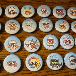 May include: Collection of round, light blue pinback buttons with cartoon character designs. Each button features a unique character with large eyes and various accessories. The buttons are arranged on a wooden surface.