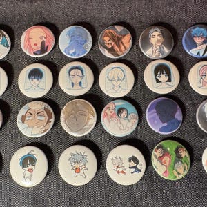 May include: A collection of 24 round, white anime-style pinback buttons. Each button features a different character illustration in various colors. The buttons are arranged on a dark gray fabric surface.