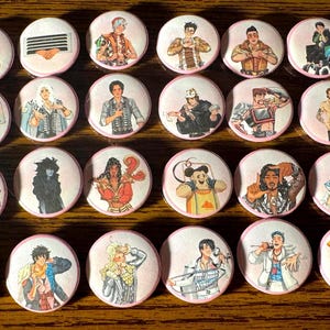 May include: A collection of round, pink-edged buttons featuring various illustrated characters. The illustrations include people in different outfits and poses, and some buttons have graphic designs. The buttons are arranged on a wooden surface.