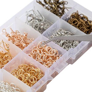 May include: A clear plastic storage container with multiple compartments filled with various metal jewelry findings, including earring hooks and jump rings in gold, silver, bronze, and rose gold tones, with a pair of metal tweezers holding a jump ring.