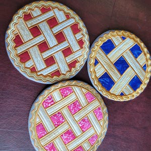May include: Three round, decorative coasters with a woven pattern. The coasters are white with a gold border and feature a red, blue, and pink woven design.
