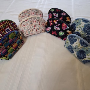 May include: A collection of various cosmetic bags with different patterns. Designs include floral, butterfly, and geometric patterns. The bags are in various colors, including blue, black, and cream. Each bag has a zipper closure.
