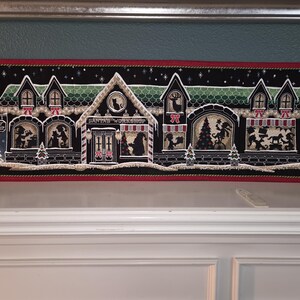 May include: A rectangular Christmas-themed wall hanging with a black background and a red border. The image depicts Santa's Workshop with silhouettes of elves, reindeer, and a Christmas tree. The text "SANTA'S WORKSHOP" is visible.