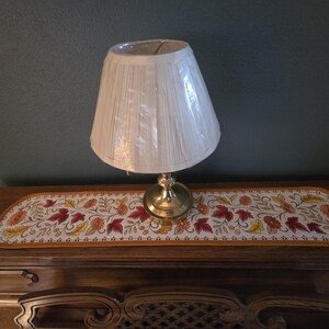 May include: A small table lamp with a cream-colored pleated lampshade sits on a wooden surface. The lamp has a brass base. A decorative runner with a floral and leaf pattern in red, orange, and yellow accents the wooden surface.