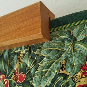 May include: A wooden hanging system for quilts or tapestries. The system is made of a solid piece of wood with a groove on the top to hold the fabric. The fabric is a green and gold floral print with red flowers.