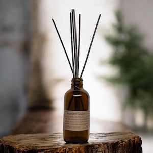 May include: A brown glass diffuser bottle with black reeds. The label reads "Marion Lake" and lists scent notes. The bottle sits on a wooden surface, with a blurred background.