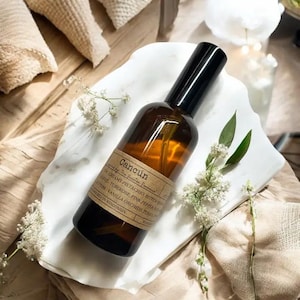 May include: A brown glass bottle with a black spray top and a label that reads "Cancún" and lists the scent notes: "Creamy Pistachio, Bitter Orange, Tuberose, Pink Pepper, Cotton Vanilla Orchids, Teakwood". The bottle is sitting on a white surface.