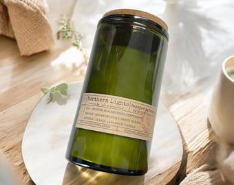 May include: A green glass candle jar with a cork lid. The jar has a label that reads "Northern Lights" with scent notes. The candle sits on a white and brown surface. The candle is hand poured in Wisconsin.