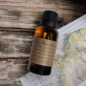May include: A brown glass bottle with a black cap and a label that reads "Rocky Mountain." The label lists the scent notes: White Pine, Mountain, Wild Huckleberry, Birchwood, and Fir. The bottle is on a wooden surface with a map.