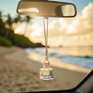 May include: A clear, square-shaped car air freshener with a wooden top and a beige string, hanging from a car's rearview mirror. The air freshener contains a light yellow liquid. The background shows a beach with palm trees and the ocean.