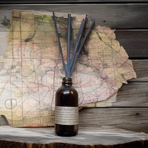 May include: A brown glass diffuser bottle with dark gray reeds, labeled "Baileys Harbor Door County". The bottle sits on a wooden surface in front of a vintage map, creating a rustic aesthetic. The label lists scents like "Lake Breeze" and "Sandalwood".