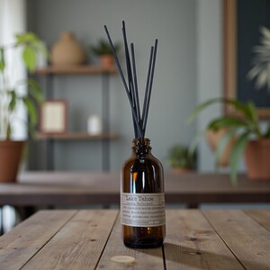 May include: A brown glass diffuser bottle with black reeds. The label reads "Lake Tahoe" and lists ingredients. The bottle sits on a wooden table, with plants and a shelf in the background. A home fragrance product.
