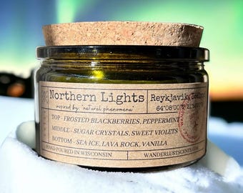 May include: A small, round glass jar with a cork lid, labeled "Northern Lights." The label lists scent notes: frosted blackberries, peppermint, sugar crystals, sweet violets, sea ice, lava rock, and vanilla. The jar is hand-poured in Wisconsin.