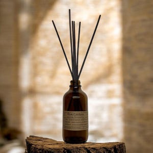 May include: A brown glass diffuser bottle with black reeds. The bottle has a label that reads "Charleston". The diffuser is placed on a wooden surface. The background is a neutral color.