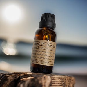 May include: A brown glass bottle with a black cap, labeled "Amalfi Coast" with scent notes. The bottle is on a wooden surface, with a blurred ocean and sky background. The label lists the scent's top, middle, and bottom notes.