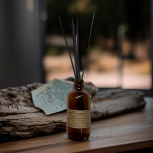 May include: A brown glass bottle with a label reading "Rocky Mountains" holds black reeds. The bottle sits on a wooden surface, with a blurred background. The reeds are arranged to diffuse fragrance.