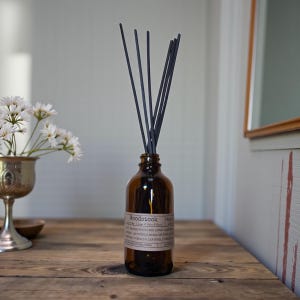 May include: A brown glass diffuser bottle with black reeds. The bottle has a label that reads "Woodstock". The diffuser is on a wooden table, with a silver vase of white flowers to the left. The background is a light gray wall.