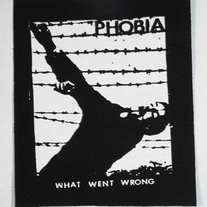 Könnte beinhalten: Schwarz-weiß-Patch mit einer Silhouette einer Person, die über einen Stacheldrahtzaun greift. Der Text "PHOBIA" befindet sich oben und "WHAT WENT WRONG" unten.