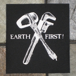 May include: Black patch with a white image of a crossed hammer and wrench. The text "EARTH FIRST!" is printed below the tools.