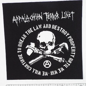 Könnte beinhalten: Schwarz-weißes Patch mit dem Text "Appalachian Terror Unit" und einem Totenkopf mit gekreuzten Knochen mit dem Text "We Win - We Will Continue To Break The Law And Destroy Property Until We Win" in einem Kreis darum.