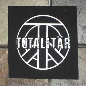 Pode incluir: Patch quadrado preto com um design de símbolo de paz branco e a palavra "TOTALITÄR" em uma fonte desgastada. O patch tem aproximadamente 7.6 cm de largura. O patch é feito de um tecido tecido.