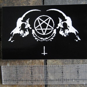 Könnte beinhalten: Schwarz-weißer Aufkleber mit einem Pentagramm, zwei Schädeln und einem umgekehrten Kreuz. Der Aufkleber ist auf schwarzem Hintergrund.